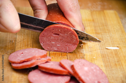 A female hand holds a knife and she cuts sausage on a wooden board. Pizza  sausage