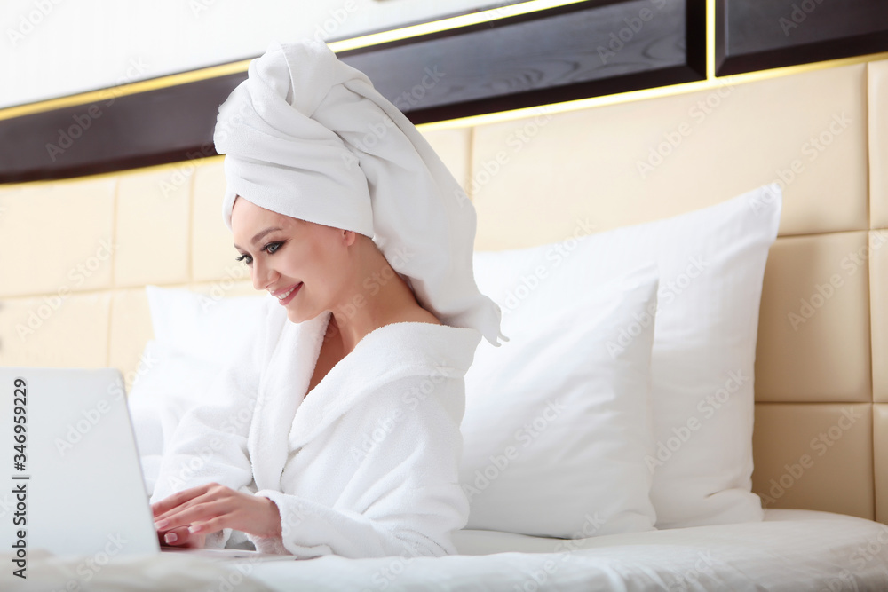 A young girl on the bed in a Bathrobe and a towel on her head is smiling and working on a laptop. The concept of distance learning or remote work. Home environment. Copy space. A young girl on the bed in a Bathrobe and a towel on her head is smiling and working on a laptop. The concept of distance learning or remote work. Home environment. Copy space.
