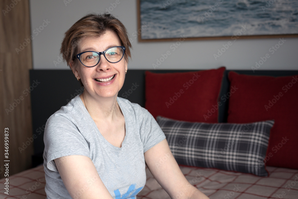Portrait of a smiling, happy middle-aged woman. She is sitting on the bed in the bedroom, at home. Portrait of a smiling, happy middle-aged woman. She is sitting on the bed in the bedroom, at home.