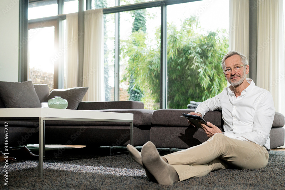Senior man with grey hair in modern design living room sitting on floor holding tablet Senior man with grey hair in modern design living room sitting on floor holding tablet