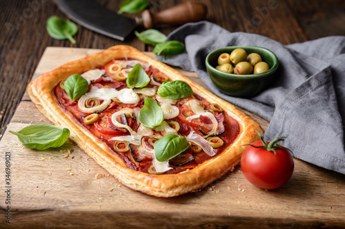 Homemade puff pastry pizza with bacon slices, tomatoes, green olives, cheese and onion, topped with basil leaves