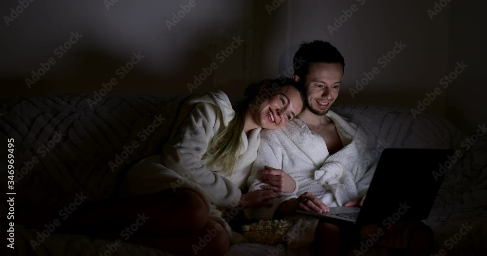 Young couple laugh and smile using computer laptop at modern home at night. Watching movie on the internet together while lying on a sofa in living room at house concept. Young couple laugh and smile using computer laptop at modern home at night. Watching movie on the internet together while lying on a sofa in living room at house concept.