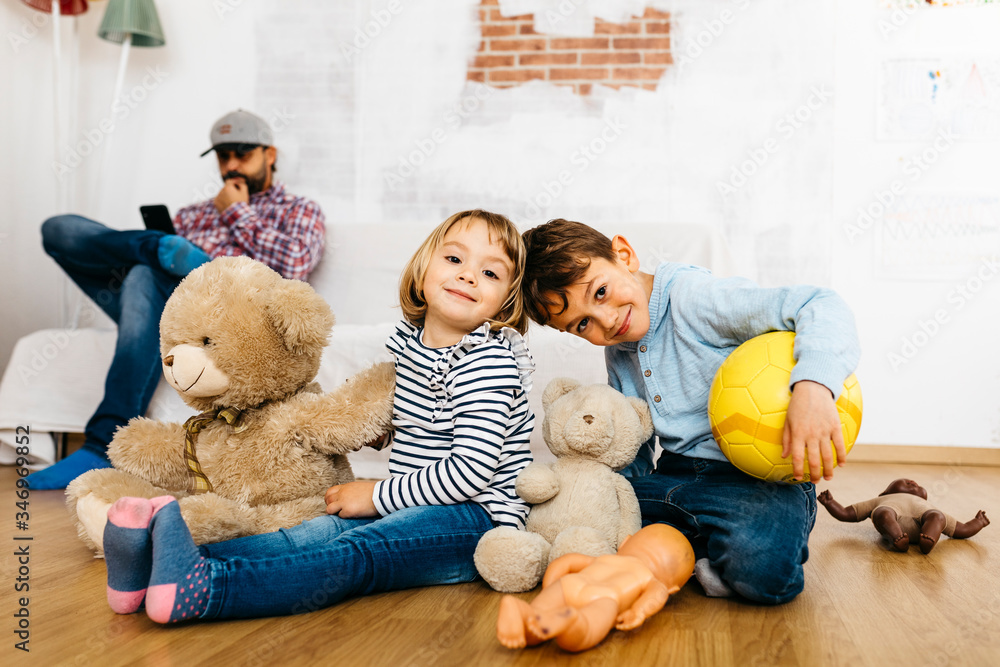 Brother and sister sitting in playroom with cuddly toys, father in background Brother and sister sitting in playroom with cuddly toys, father in background