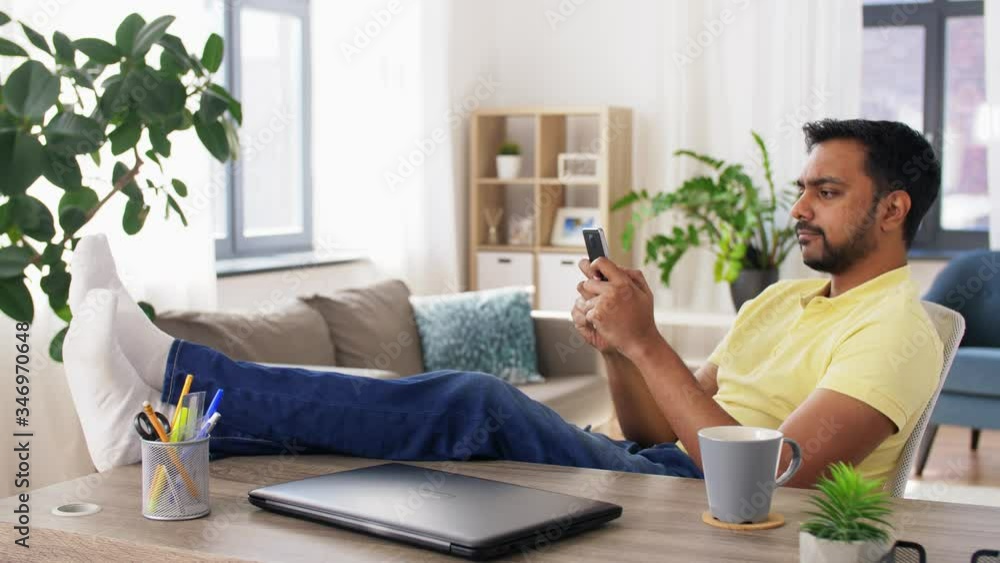 technology, remote job and people concept - indian man with smartphone resting feet on table at home office technology, remote job and people concept - indian man with smartphone resting feet on table at home office