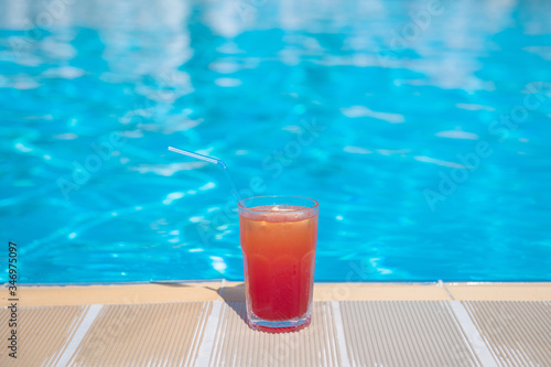 Fresh alcoholic red cocktail sex on beach with ice in glass on edge of pool in summer weather against background washed up pool and recreation area. Concept good holiday and travel, place for text.