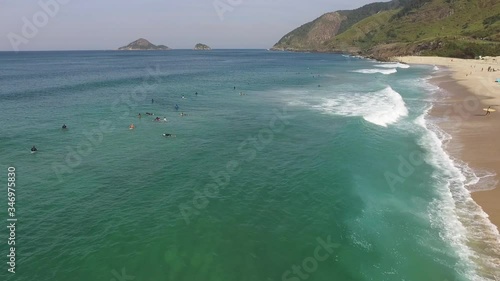 Surfers in a calm sea with small waves and waiting for a bigger one in a cristaline blue water in a beach with a green mountain on back. Drone aerial footage. Rio de Janeiro Brazil photo