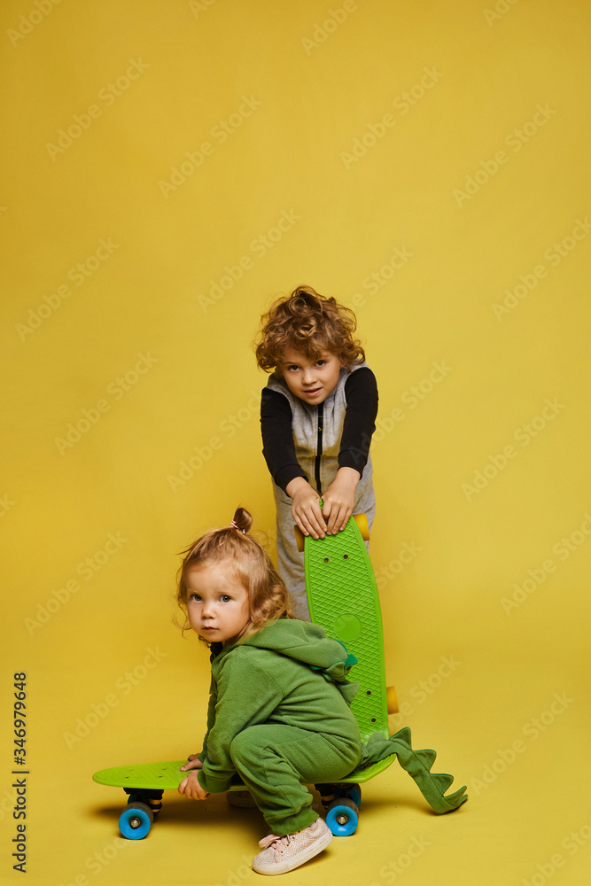 Stylish children in modish outfits, little girl and little boy in overalls posing with skateboards at the yellow background, isolated. Kids fashion. Street sport Stylish children in modish outfits, little girl and little boy in overalls posing with skateboards at the yellow background, isolated. Kids fashion. Street sport