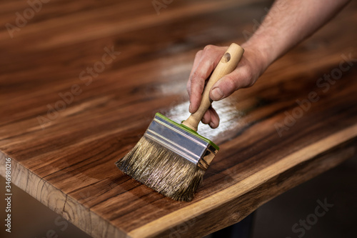 Hand of a carpenter designer oiling an authentic self made table with a big brush in a workshop environment. Finishing beautiful teak wood slabs with protective oil