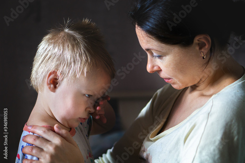 Tired mother hugging and calming little cute sad upset crying caucasian blond toddler boy at home indoor. Mom holding sleepy sick son. Child health, love and care concept.