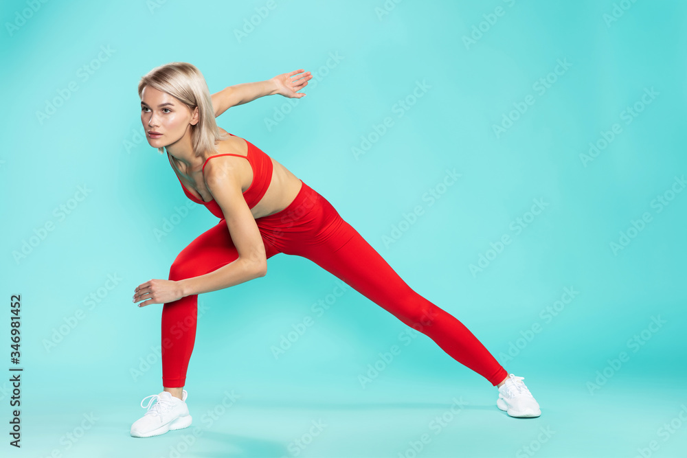 Fitness girl. Full length of young and sporty blonde woman in red sportswear exercising against blue background. Studio shot. Sport concept