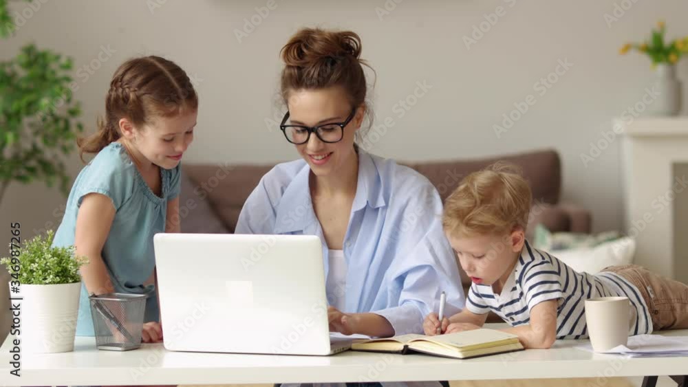 Young female in glasses doing remote project on laptop and communicating with kids while working at home Young female in glasses doing remote project on laptop and communicating with kids while working at home