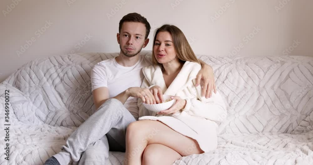 Young couple watching TV on the grey sofa at home in bright living room. Exciting couple interested on tv show eating popcorn Young couple watching TV on the grey sofa at home in bright living room. Exciting couple interested on tv show eating popcorn