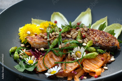 Selective focus wild herbs salad with grilled salmon, carrot, wild garlic buds, roasted sunflower kernels,chicory and  maple leaves . Garnished with edible wild flowers, dandelion and daisy flowers.