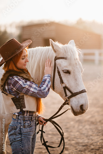 A young cowgirl woman stroking a horse in the sunset at her ranch. Flare effect.