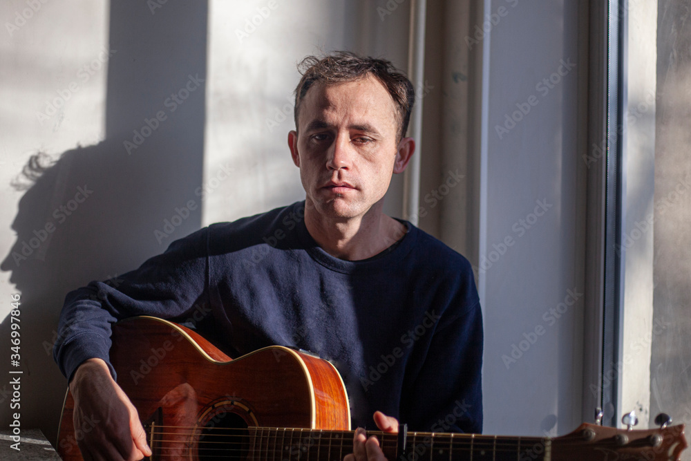 Obraz premium Guy with a guitar in the kitchen. Natural light comes to the musician from the window. Lesson playing the guitar. Unknown talented musician. Playing an acoustic guitar. Brutal man shows skill.
