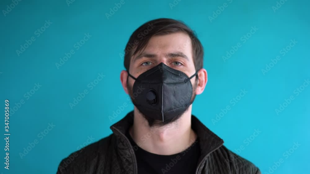 Gorgeous young bearded man with freckles with short brown hair in black jacket, t-shirt wearing protective face mask on a blue wall background, looking straight. Protection concept in lockdown period. Gorgeous young bearded man with freckles with short brown hair in black jacket, t-shirt wearing protective face mask on a blue wall background, looking straight. Protection concept in lockdown period.