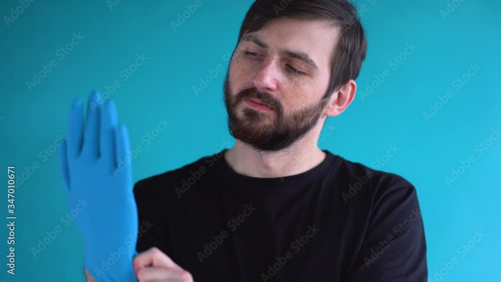 Young bearded man with freckles with short brown hair in black t-shirt put on two protective medical gloves on his hands against blue wall background close up. Protection concept in pandemic period. Young bearded man with freckles with short brown hair in black t-shirt put on two protective medical gloves on his hands against blue wall background close up. Protection concept in pandemic period.