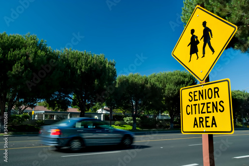 Yellow traffic sign that says Senior Citizens Area with graphic of senior citizens walking . Car is blurred driving by in background