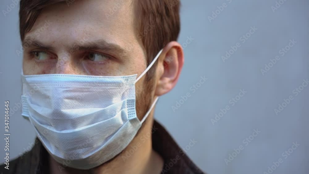 Sick infected bearded man with green eyes wear protective disposable medical face mask, stares longingly at the camera close-up. Portrait of ill beautiful guy on gray background. Isolation concept. Sick infected bearded man with green eyes wear protective disposable medical face mask, stares longingly at the camera close-up. Portrait of ill beautiful guy on gray background. Isolation concept.