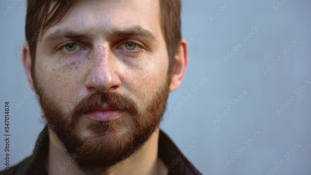 Handsome young Caucasian man with thick beard, freckles in black shirt with brown hair looks directly at camera on gray wall background. Portrait of fine guy outdoor on sunny day close up slow motion. Handsome young Caucasian man with thick beard, freckles in black shirt with brown hair looks directly at camera on gray wall background. Portrait of fine guy outdoor on sunny day close up slow motion.