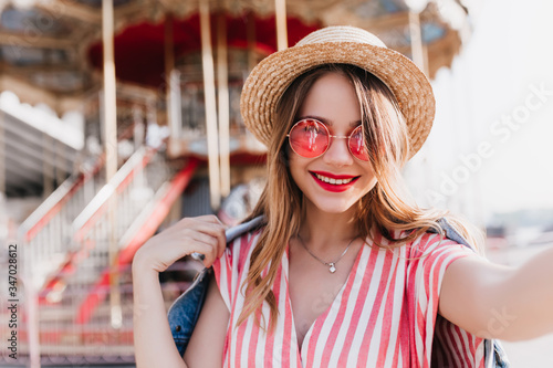 Refined young lady in elegant hat making selfie in front of carousel. Outdoor portrait of debonair caucasian girl spending weekend in amusement park.