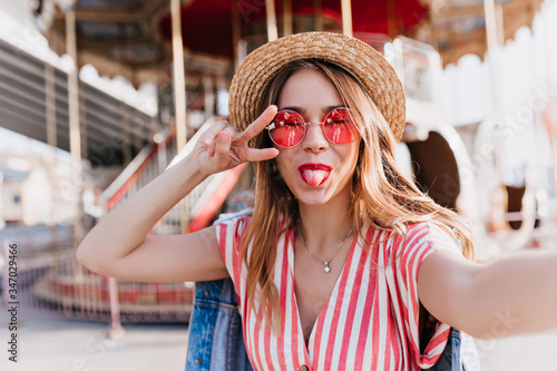 Картината върху платно Enthusiastic cute girl in pink sunglasses posing in amusement park with tongue out