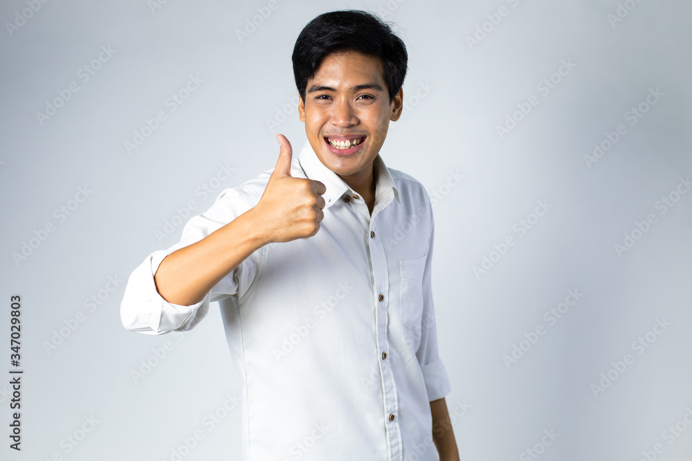 Happy handsome Asian man in white shirt showing thumbs up on gray background