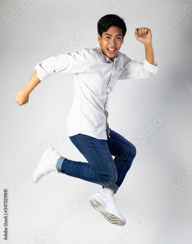 portrait of an excited young Asian man in white shirt jumping while celebrating success on gray background.