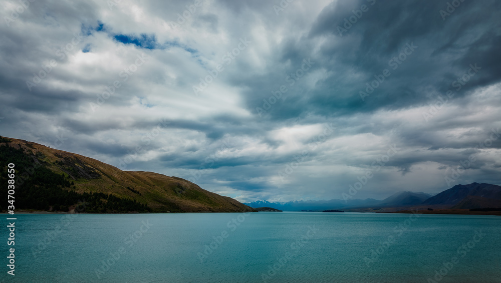 Dramatic view of Lake Pukaki with clouds gathering over the snow-capped mountain peaks. The alpine lake is famous for the amazing turquoise hues of the water and great mountain ranges in New Zealand.