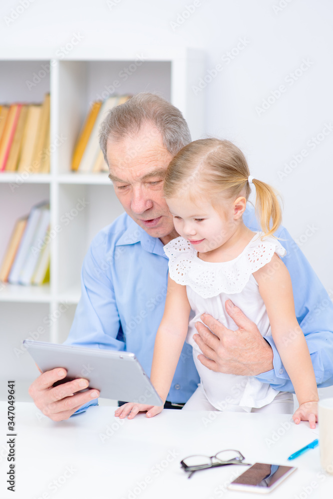 An elderly man and a little girl sitting on his lap are sitting in a room. Grandfather holding a tablet in his hand and looking at granddaughter An elderly man and a little girl sitting on his lap are sitting in a room. Grandfather holding a tablet in his hand and looking at granddaughter