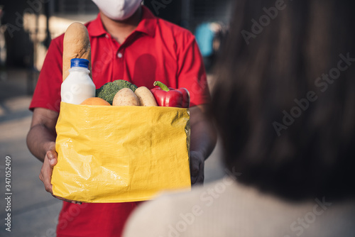 Deliver man wearing face mask in red uniform handling yellow bag of food, fruit, milk, vegetable give to female costumer Postman and express grocery delivery service during covid19.