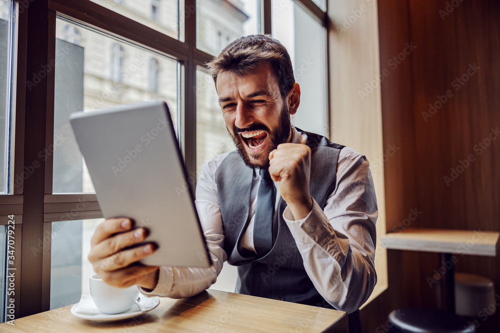 Fototapeta premium Young successful businessman sitting in coffee shop, looking at tablet and celebrating.