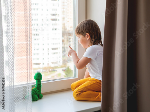 Toddler boy sits with green dragon or dinosaur. Kid with plush toy look through window outside. Child on home quarantine because of coronavirus COVID-19 waiting for somebody. photo