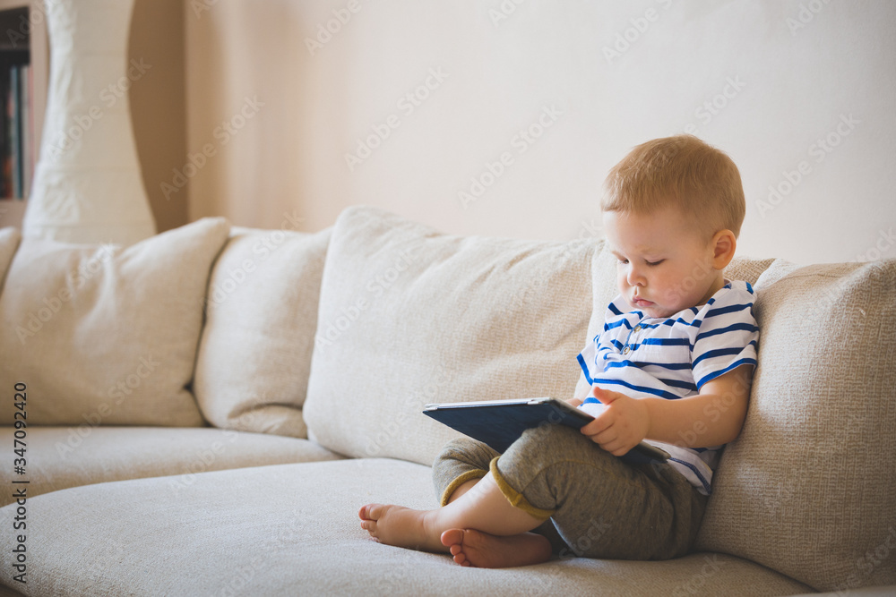 Cute boy working on a tablet while sitting on a sofa. A child plays educational games on a laptop computer at home. Early development for children. Toning and artificial noise Cute boy working on a tablet while sitting on a sofa. A child plays educational games on a laptop computer at home. Early development for children. Toning and artificial noise