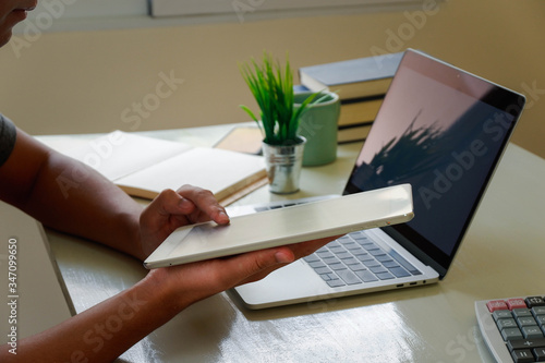 A man uses a laptop at home while sitting at a wooden table. Man hands typing on a notebook keyboard. The concept of young people working at mobile devices. The background is wide blurry window.