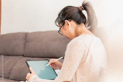 Young Asian modern woman working in graphic design and art from home. Girl sitting and drawing on an ipad tablet on the sofa during coronavirus pandemic quarantine.