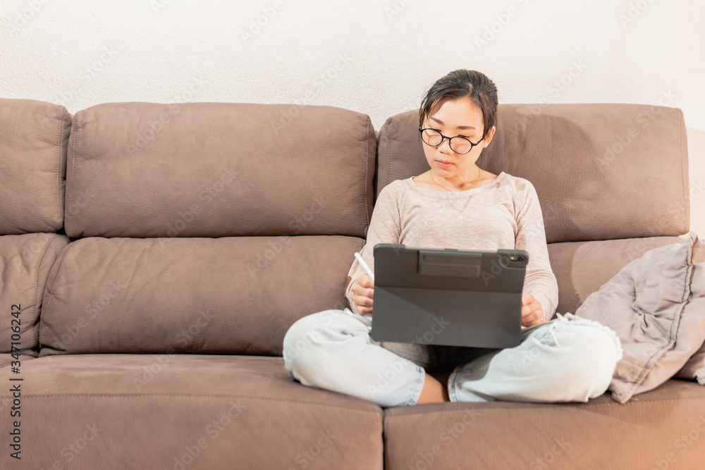 Young asian woman graphic designer drawing on an ipad tablet. Girl working from home sitting on the sofa couch. Work from home due to coronavirus pandemic. Young asian woman graphic designer drawing on an ipad tablet. Girl working from home sitting on the sofa couch. Work from home due to coronavirus pandemic.