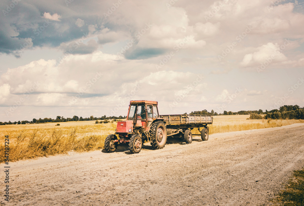 Old Red Tractor with a Trailer moving on the Countryside Sandy Road in hot summer day. Father and Son inside a tractor. Old Red Tractor with a Trailer moving on the Countryside Sandy Road in hot summer day. Father and Son inside a tractor.