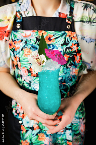 Lady bartenger holding a blue hawaii cocktail in a hurricane glass garnished with a banana leaf, flower and pink umbrella