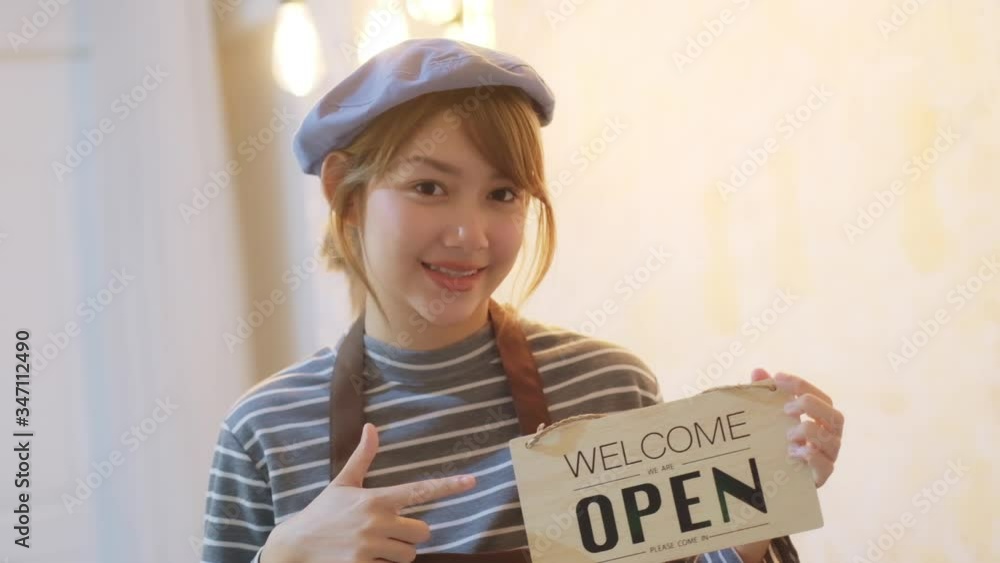 Business owner attractive young Asian woman in apron hanging we're open sign on front door smiling welcoming clients to new cafe. Beautiful Young Cafe Owner Turning Storefront Sign From Close to Open. Business owner attractive young Asian woman in apron hanging we're open sign on front door smiling welcoming clients to new cafe. Beautiful Young Cafe Owner Turning Storefront Sign From Close to Open.