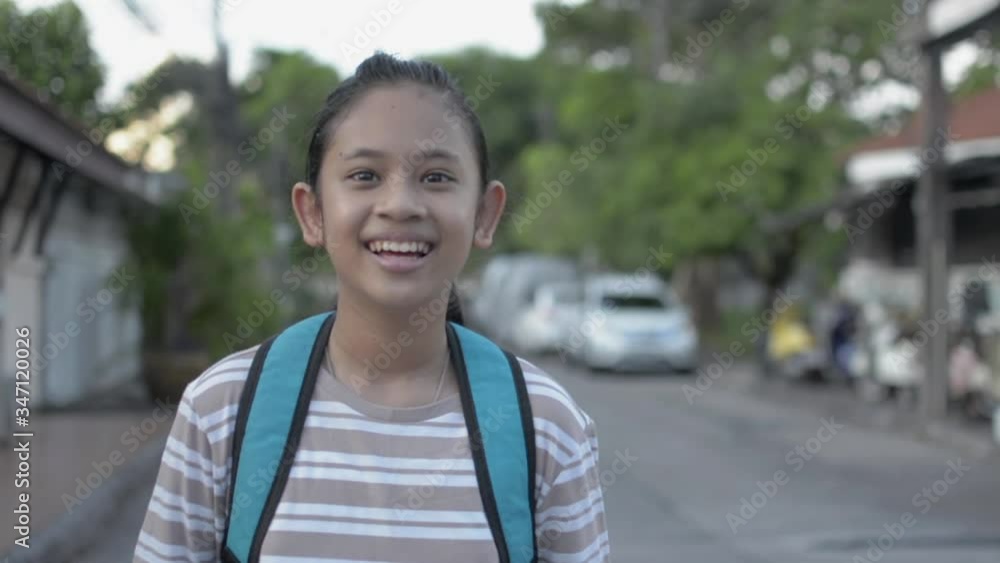 Portrait of cute asian girl in casual dress with backpack is smiling and talking during standing on the footpath in the housing area. Pretty school girl looking at camera and laughing on the road. Portrait of cute asian girl in casual dress with backpack is smiling and talking during standing on the footpath in the housing area. Pretty school girl looking at camera and laughing on the road.