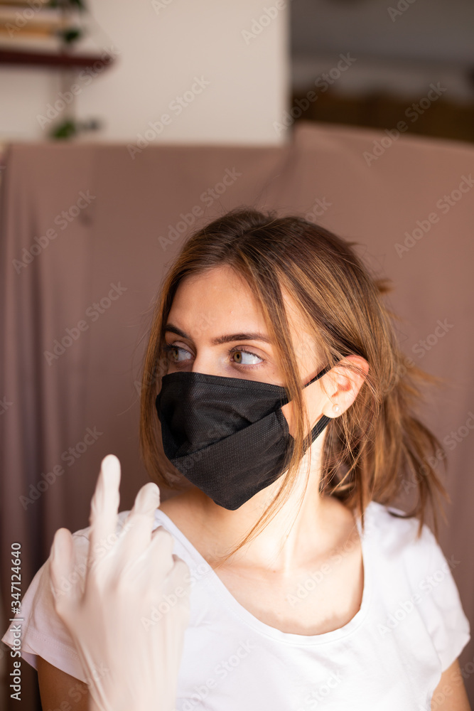 portrait of beautiful young woman wearing cotton black mask and white medical/surgical gloves on pastel background. She is adjusting mask. portrait of beautiful young woman wearing cotton black mask and white medical/surgical gloves on pastel background. She is adjusting mask.