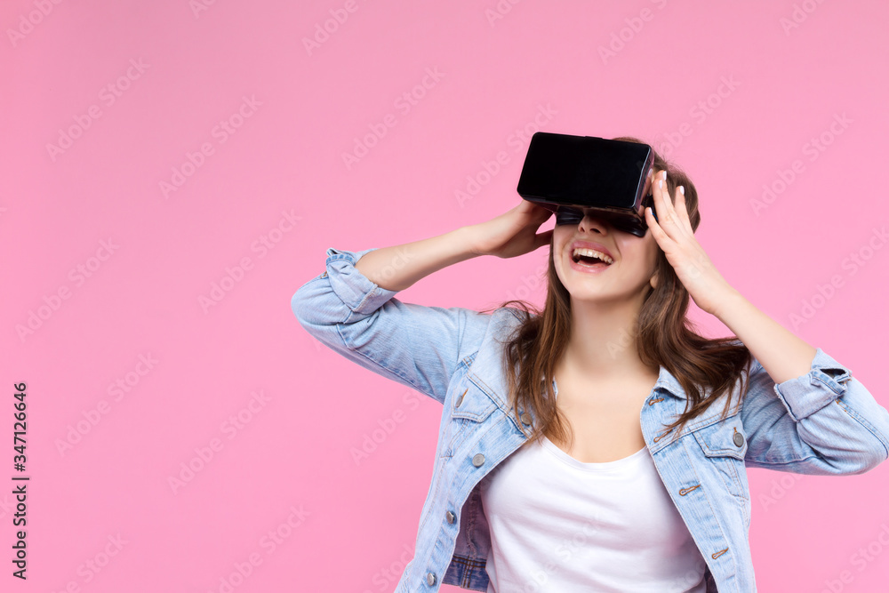 Pretty, smiling, surprised girl in VR glasses on pink background. Young woman dressed in denim jacket, white t-shirt is enjoying virtual reality world. Modern technology and devices concept. Pretty, smiling, surprised girl in VR glasses on pink background. Young woman dressed in denim jacket, white t-shirt is enjoying virtual reality world. Modern technology and devices concept.