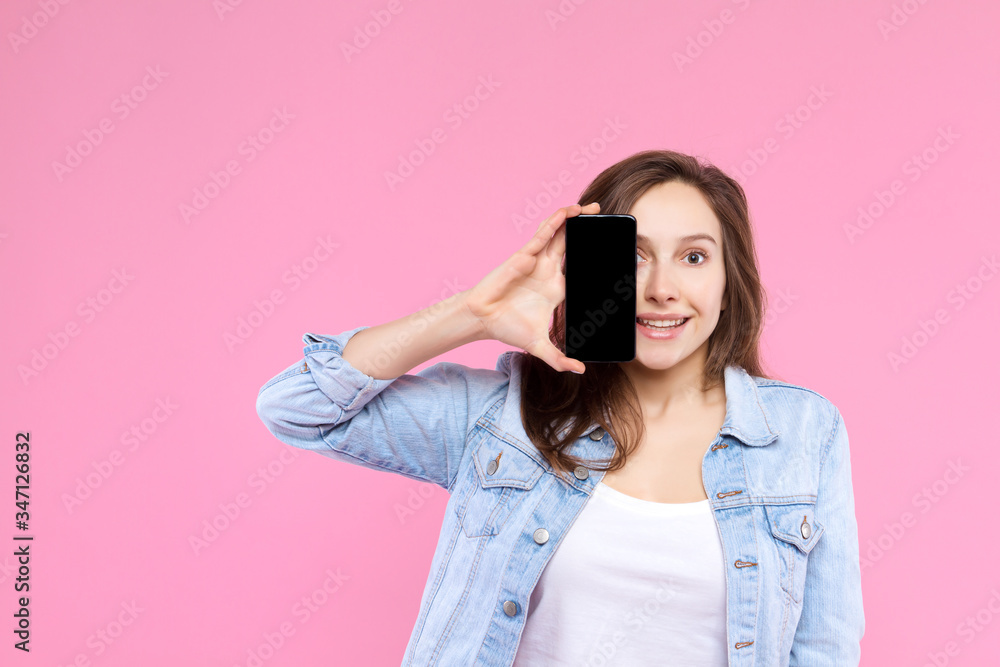 Pretty smiling girl is holding in hand smartphone with empty blank screen display. Young woman dressed in denim jacket and white t-shirt on pink background. Modern technology and devices concept. Pretty smiling girl is holding in hand smartphone with empty blank screen display. Young woman dressed in denim jacket and white t-shirt on pink background. Modern technology and devices concept.
