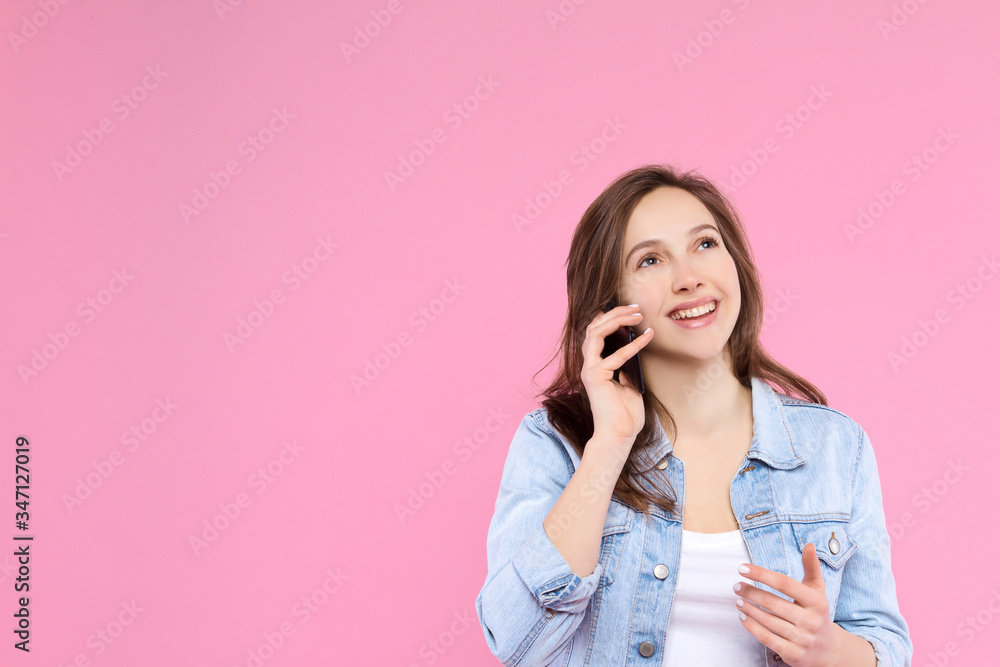 Pretty, smiling girl is holding in hand smartphone and talking. Young woman dressed in denim jacket and white t-shirt on pink background. Modern technology and devices concept. Mobile connection. Pretty, smiling girl is holding in hand smartphone and talking. Young woman dressed in denim jacket and white t-shirt on pink background. Modern technology and devices concept. Mobile connection.