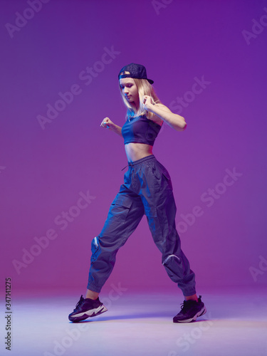 Cute teen girl dancing hip-hop in stylish clothes, a baseball cap, in a Studio with neon lighting. Dance color poster.