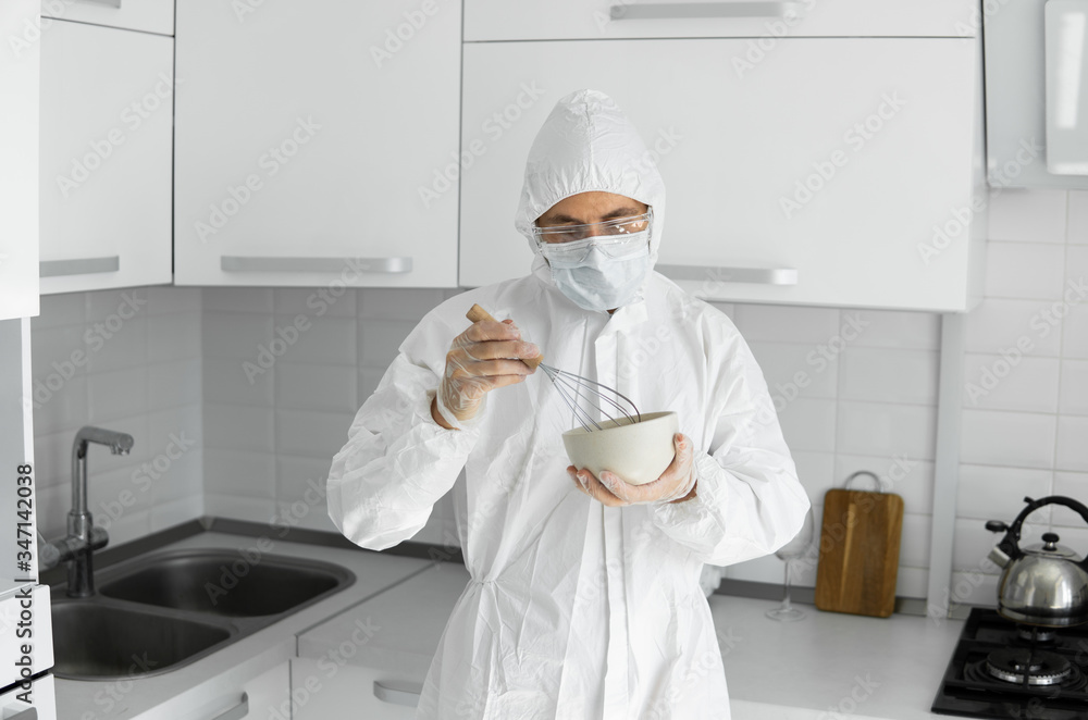 Man in white protective suit and in face medical mask cooks in the white kitchen at home during coronavirus. Stay at home. Enjoy cooking at home. COVID-19 global pandemic.