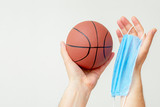 Hands of man holding basketball ball with medical mask on a light background with copy space. Coronavirus suspends world sport.