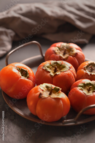 Fototapeta Naklejka Na Ścianę i Meble -  Persimmon. Orange. Ripe persimmons on a dark plate. Vintage fruit plate. Fruit. Dark background. Leaves