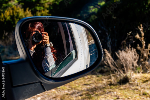 A close-up shot of the reflection of an unrecognizable young Caucasian redhead female photographer holding a DSLR photo camera in the rearview mirror of a moving car
